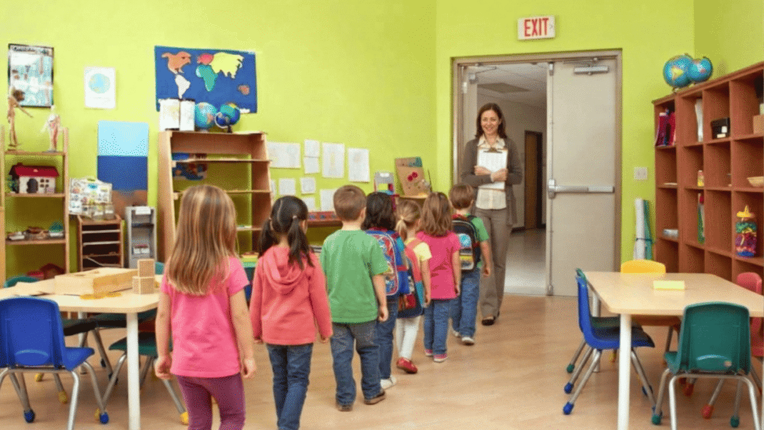 Children line up in a classroom, heading toward a teacher standing by the door. The room has bright walls, tables, chairs, and educational materials.