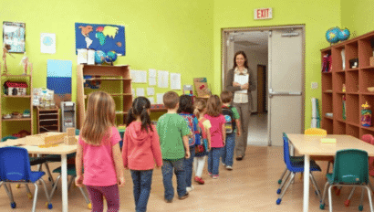 Children line up in a classroom, heading toward a teacher standing by the door. The room has bright walls, tables, chairs, and educational materials.